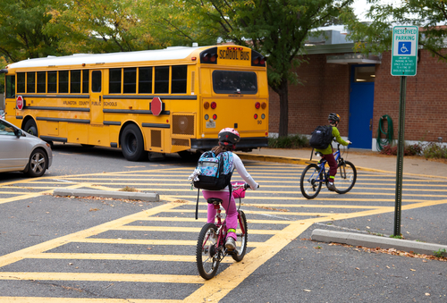 Two children riding bikes with helmets enter a school driveway marked with yellow crosswalk lines while a yellow school bus is parked next to the sidewalk. Green reserved parking sign and brick building are in the background.