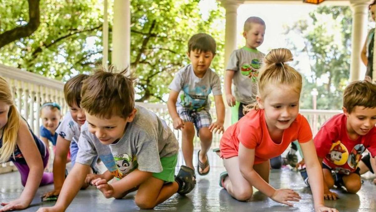 A group of eight children playfully racing on all fours across a sunny porch with a white railing, surrounded by greenery.